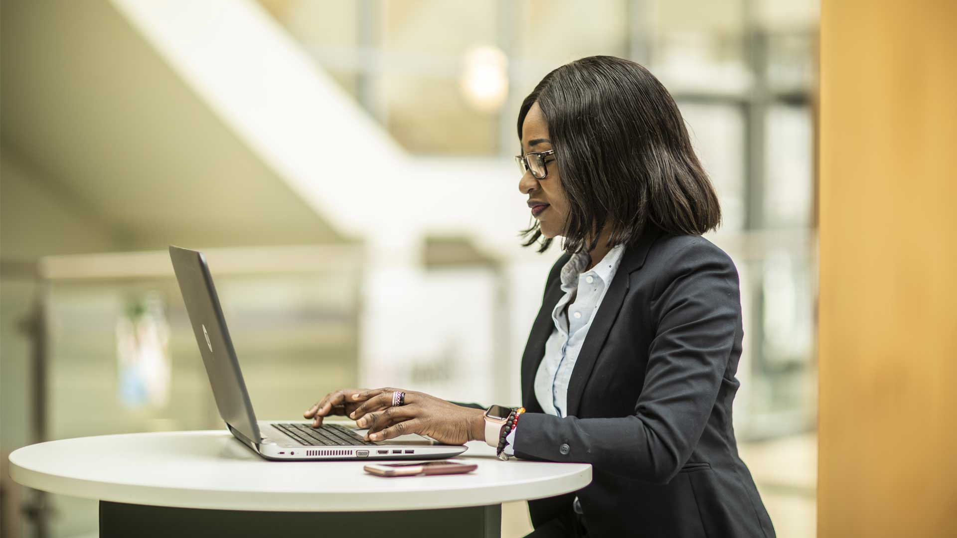 A student sits with her laptop open on the table. The background is blurred.